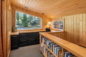A cozy cabin interior with a wooden desk, bookshelves, a lamp, and a window overlooking a forest.
