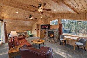 L’intérieur chaleureux d’un chalet avec une cheminée en pierre, des meubles en cuir, un logo Netflix et une grande fenêtre donnant sur une forêt.