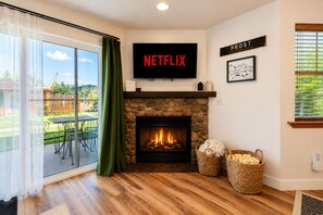 A living room with a stone fireplace, a large TV displaying 'NETFLIX,' and a view of a backyard through a sliding glass door.