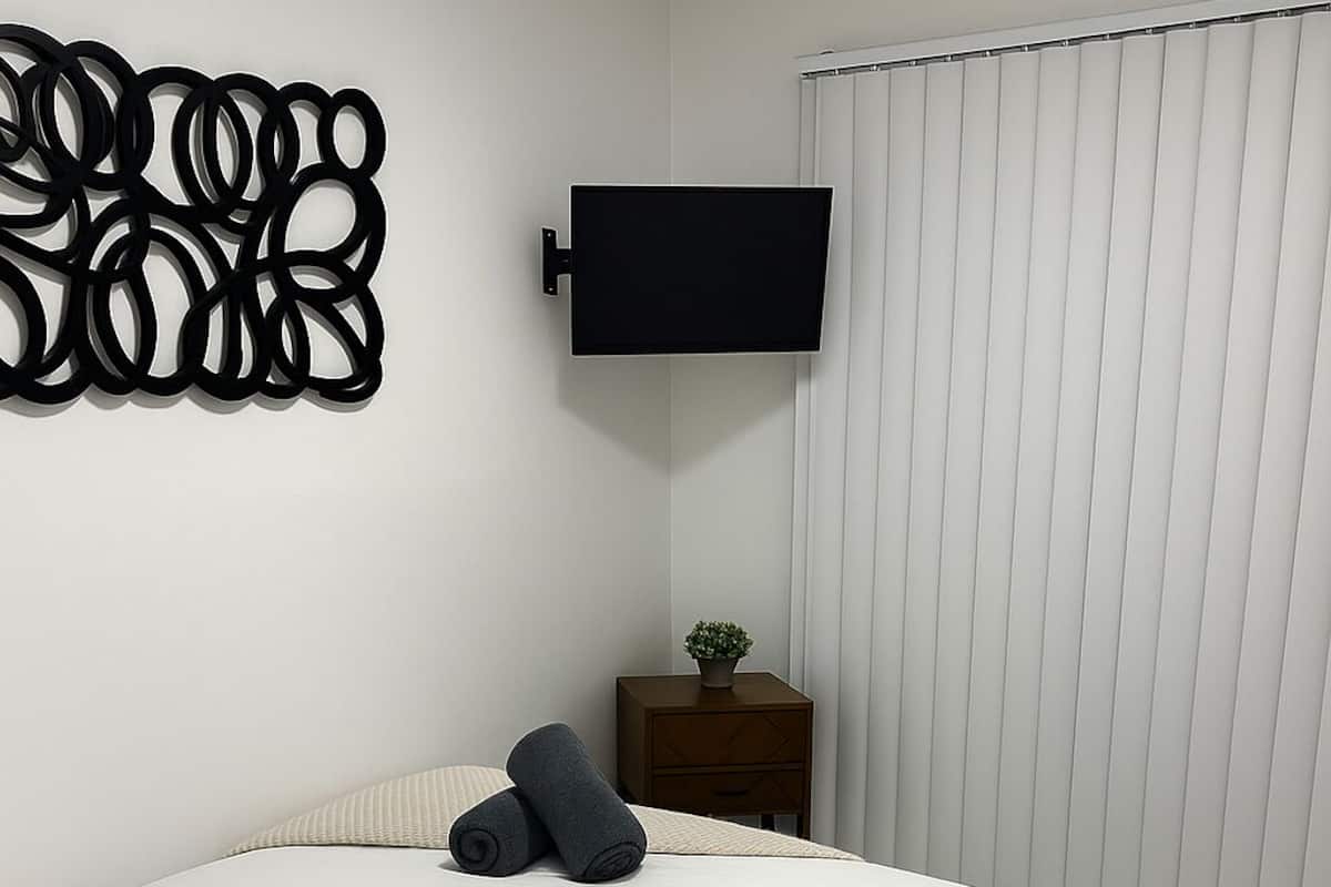 Ein ordentlich bezogenes Bett mit Kissen, ein kleiner Nachttisch mit Pflanze und ein an der Wand befestigtes Fernsehgerät.