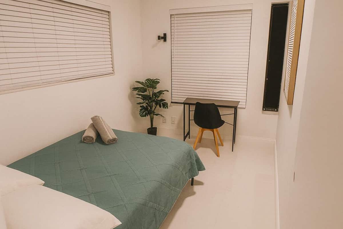 Ein Schlafzimmer mit Bett, Schreibtisch, Stuhl, Topfpflanze und einem Fenster mit Jalousien.