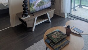 A modern hotel room with a flat-screen TV on a wooden stand, a bed with white bedding, a small round table with books and a vase, and a large window with a view of greenery and buildings.