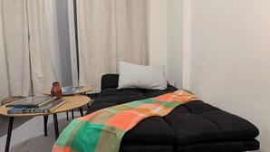 A bedroom with a black sofa bed, a small wooden table with books, a white pillow, and a checkered blanket.