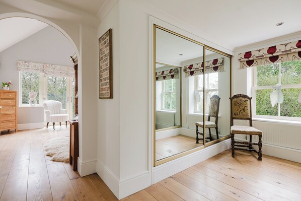 Une pièce dotée d’un grand miroir, d’un plancher en bois, de murs blancs, d’une chaise blanche, d’une commode en bois et d’une fenêtre donnant sur un paysage verdoyant.