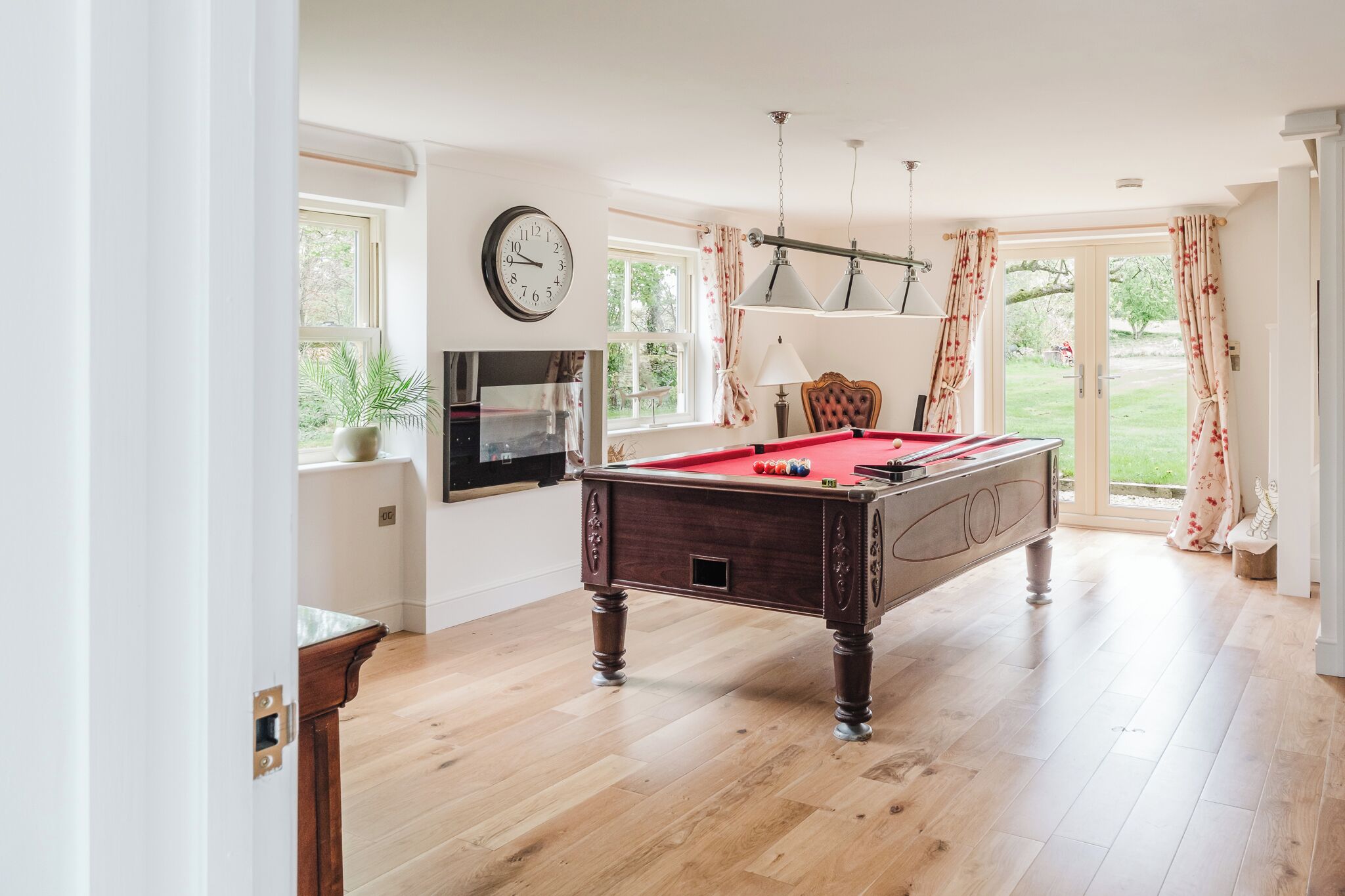 A billiards table in a room with wooden flooring, a clock on the wall, and a window with curtains.