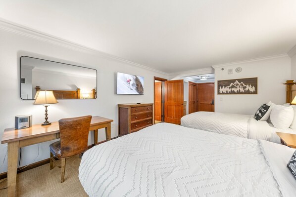 Une chambre d’hôtel comprenant un lit, un bureau avec une chaise, une commode, un miroir et deux tableaux accrochés au mur.