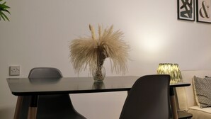 A modern dining area with a glass-top table, a vase with pampas grass, a black chair, a sofa with a patterned throw, and abstract wall art. - Spacious apartment in central London - 10-15 minutes from major attractions! (London)