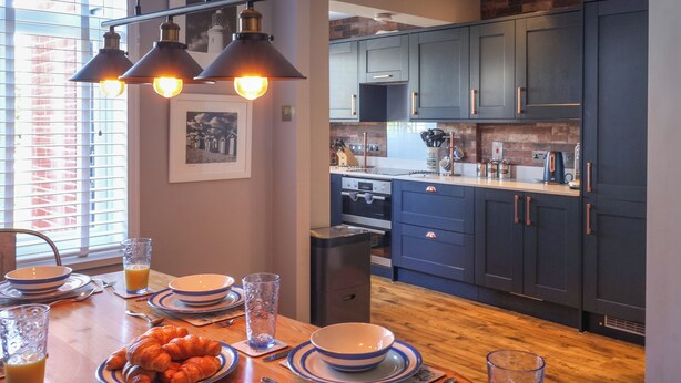 A modern kitchen with dark blue cabinets, a wooden countertop, and a dining area with a table set for breakfast. - The Waverley (Reydon)