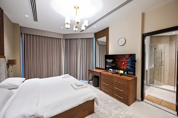 A modern hotel room with a large bed, a TV, a wooden dresser, and a glass shower.