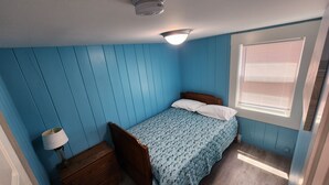 A small, single-bed room with blue walls, a wooden headboard, a bedside table with a lamp, and a window with blinds.