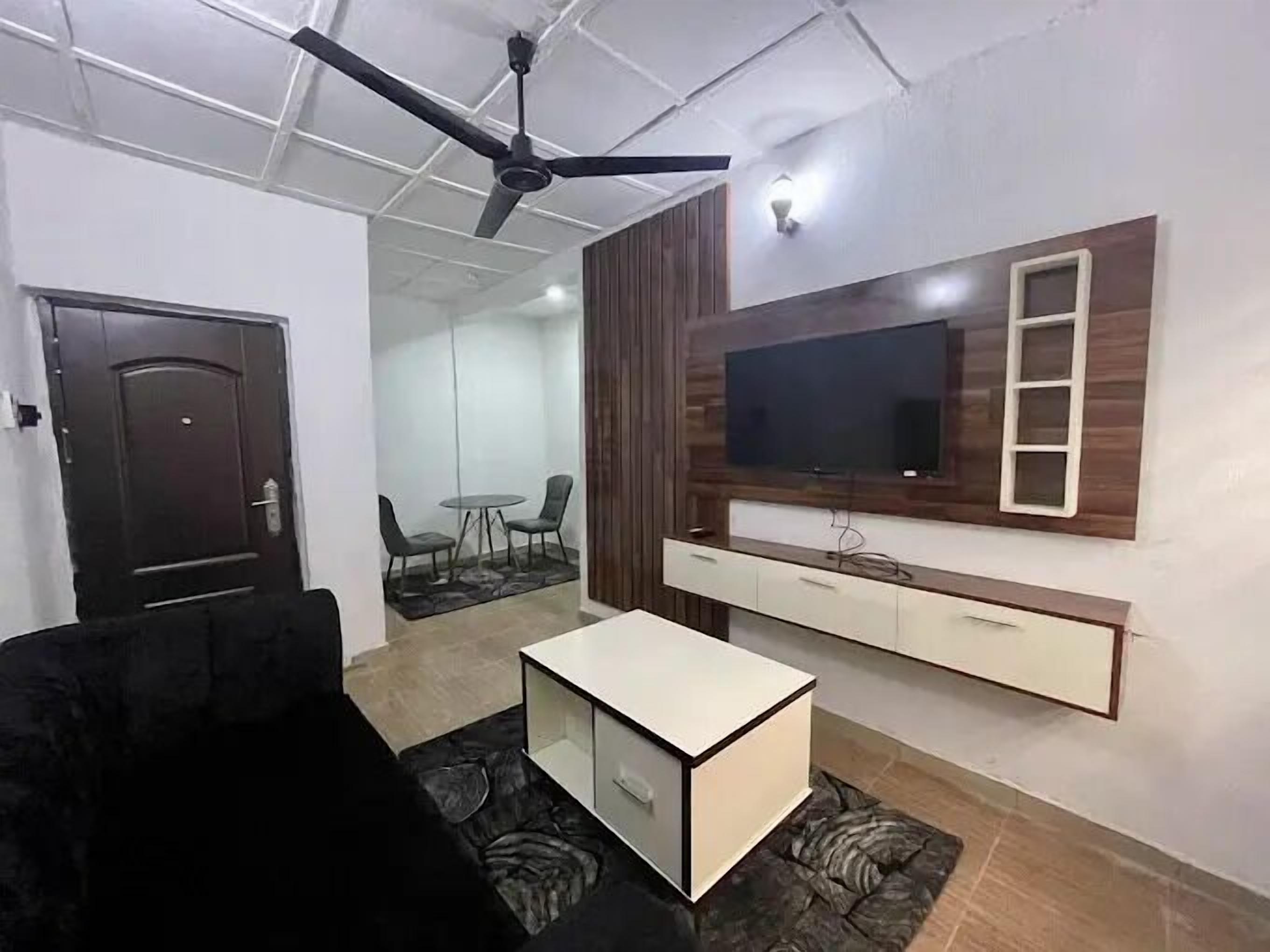 A modern living room with a ceiling fan, a flat-screen TV mounted on a wooden panel, a white cabinet below, and a black sofa.