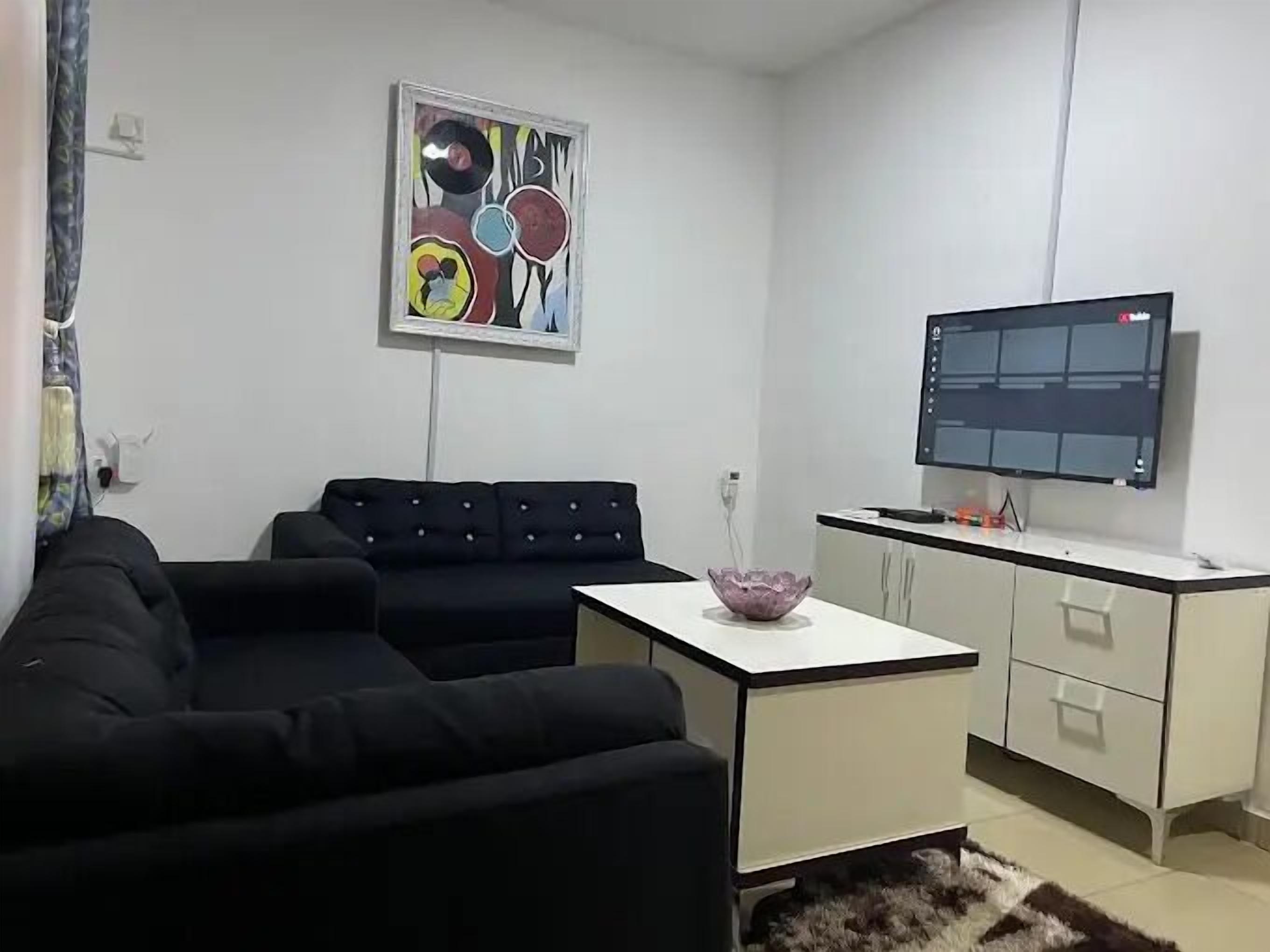 Una sala moderna con un sofá seccional negro, una mesa de centro blanca, un cuadro abstracto colorido, un televisor de pantalla plana y una zona de cocina con armarios blancos.