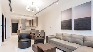 A modern living room with a sofa, armchair, and coffee table. There is a dining area with chairs and a table. The room has a large window with a view of the sea.