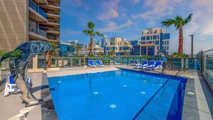 A rooftop pool with blue water, surrounded by lounge chairs and palm trees, with modern buildings in the background.