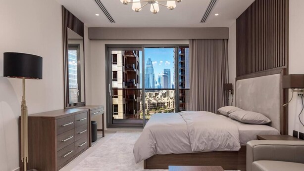 A modern bedroom with a large bed, a dresser, a TV, and a view of a cityscape through a sliding door.