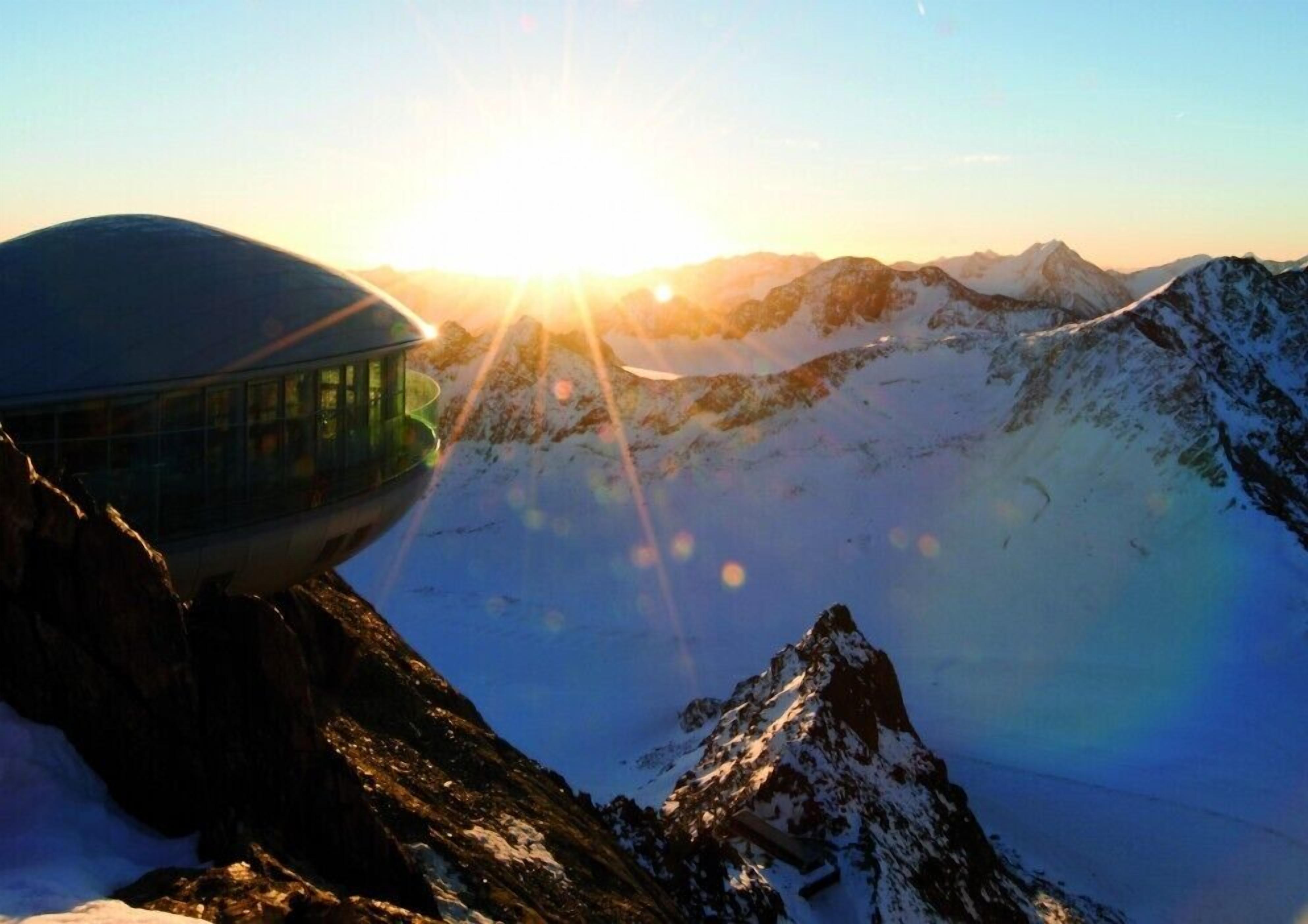 A mountain hut with a glass enclosure, surrounded by snow-covered peaks and a clear sky.