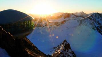 A mountain hut with a glass enclosure, surrounded by snow-covered peaks and a clear sky.