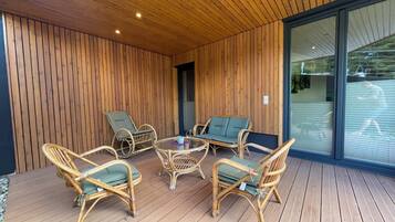 Una terraza de madera con mobiliario de mimbre y un ventanal que da a un jardín.
