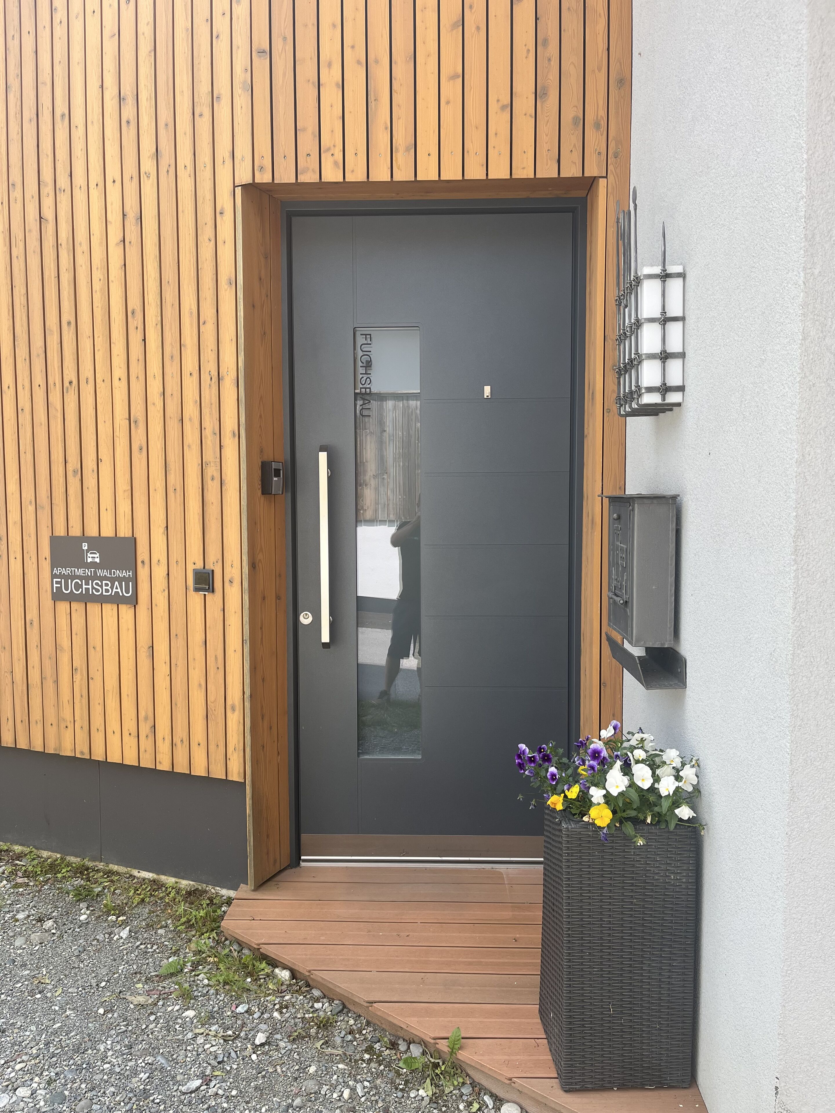 Una entrada moderna a un edificio con una puerta oscura, paneles de madera en la pared, un letrero que indica 'Apartment Waldnah Fuchsbau' y una maceta con flores.