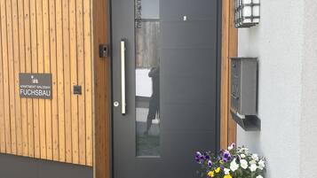 Una entrada moderna a un edificio con una puerta oscura, paneles de madera en la pared, un letrero que indica 'Apartment Waldnah Fuchsbau' y una maceta con flores.