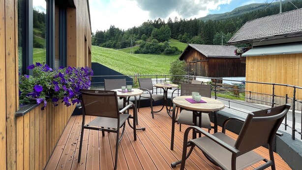 A wooden deck with outdoor seating, overlooking a green valley and a mountain range. - Apartment Waldnah (Fließ)
