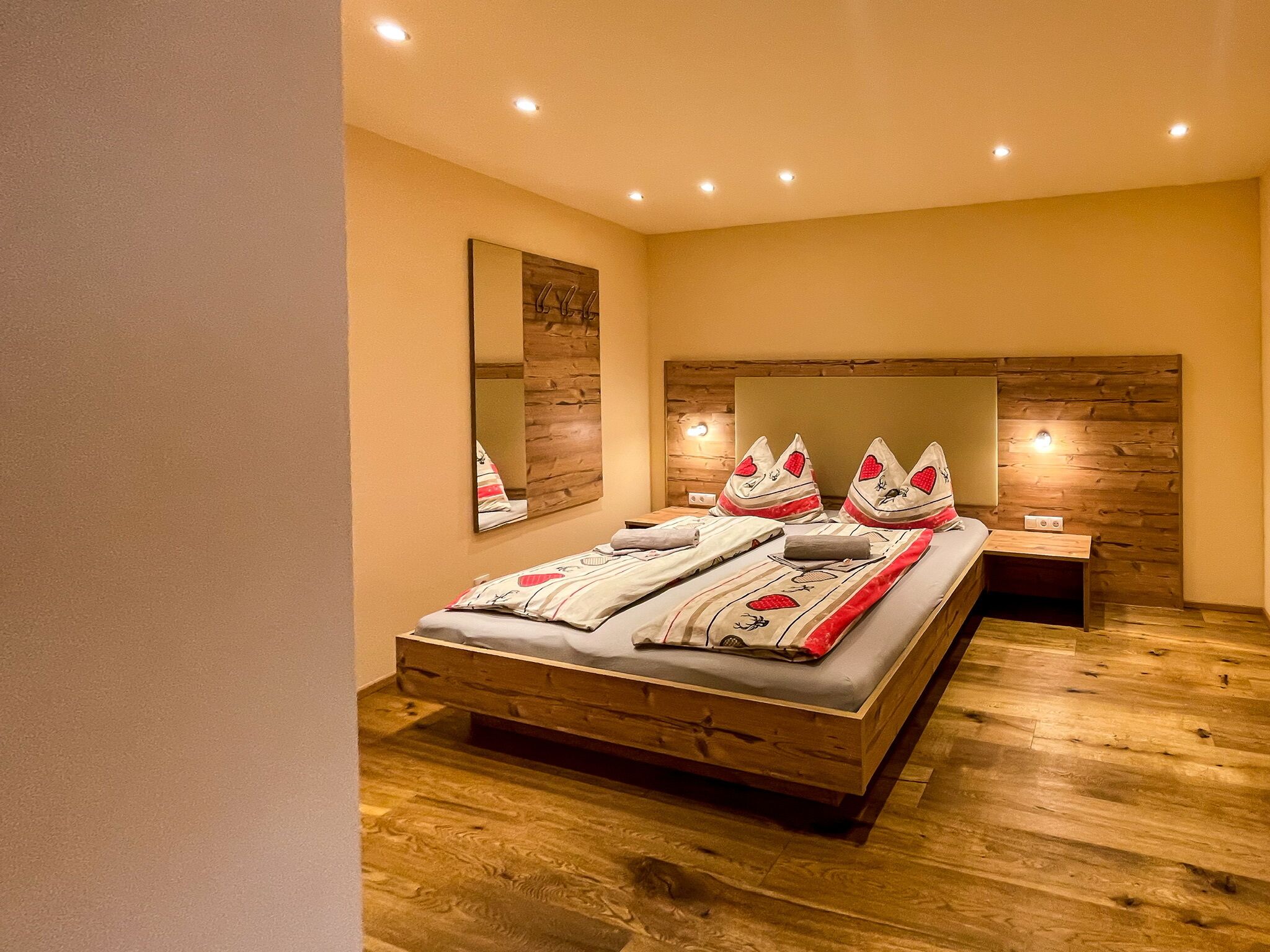 A bedroom with a wooden headboard, a bed with patterned bedding, a nightstand, and a wall-mounted mirror.