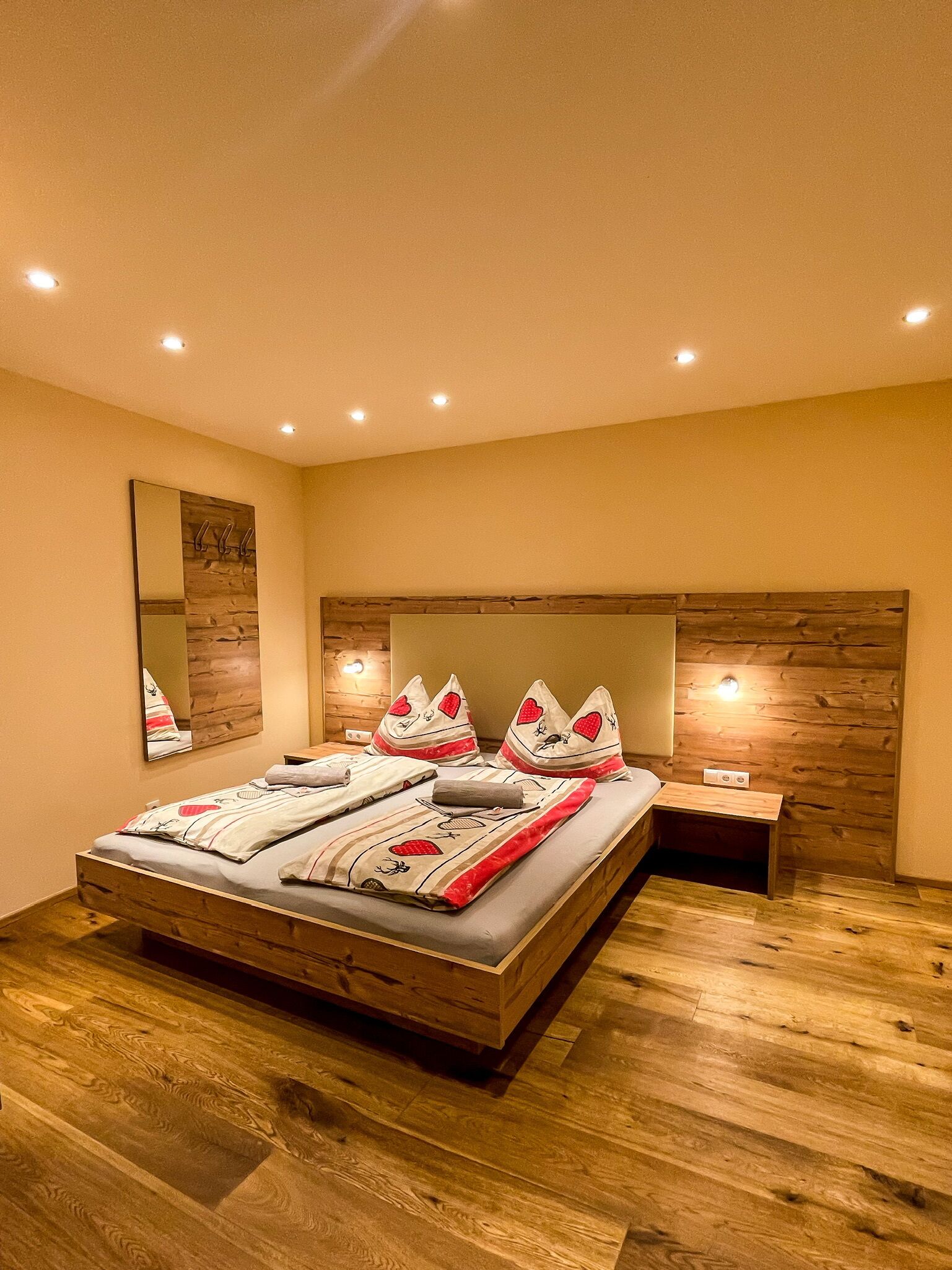 A bedroom with a wooden headboard, a large bed with striped bedding, a bedside table, and a wall-mounted mirror.