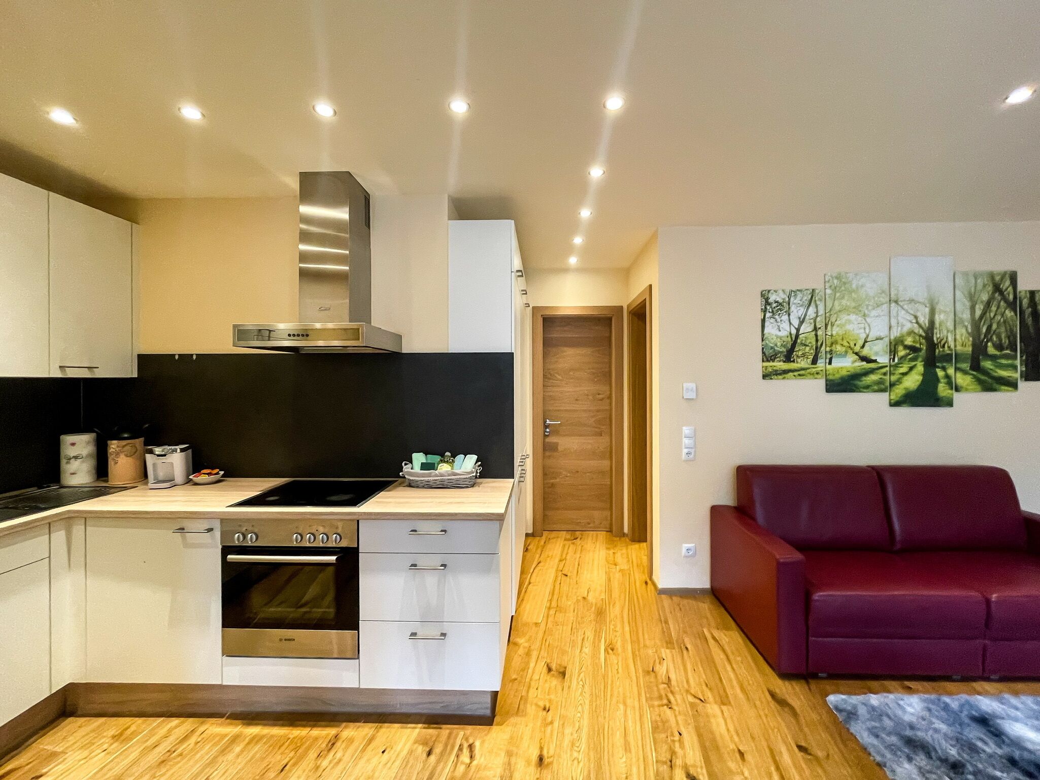A modern kitchen with a built-in oven, a stove, and a microwave. There is a wooden floor and a red sofa in the living area.