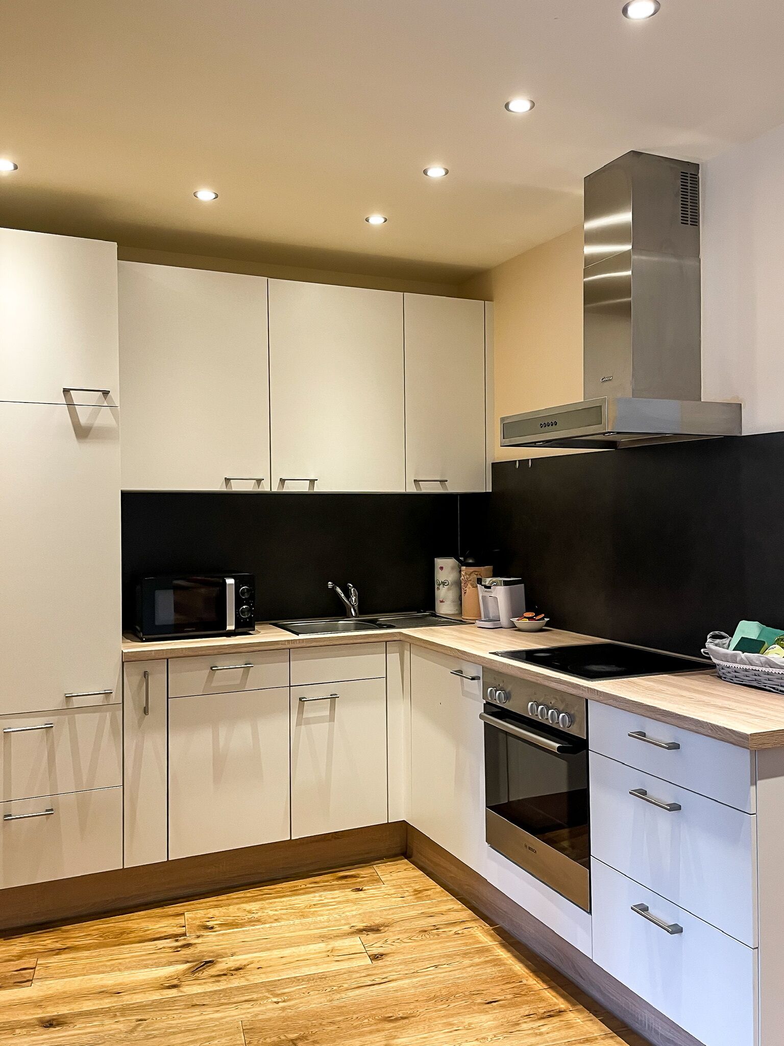 A modern kitchen with white cabinets, a black countertop, a stainless steel range hood, and a wooden floor.