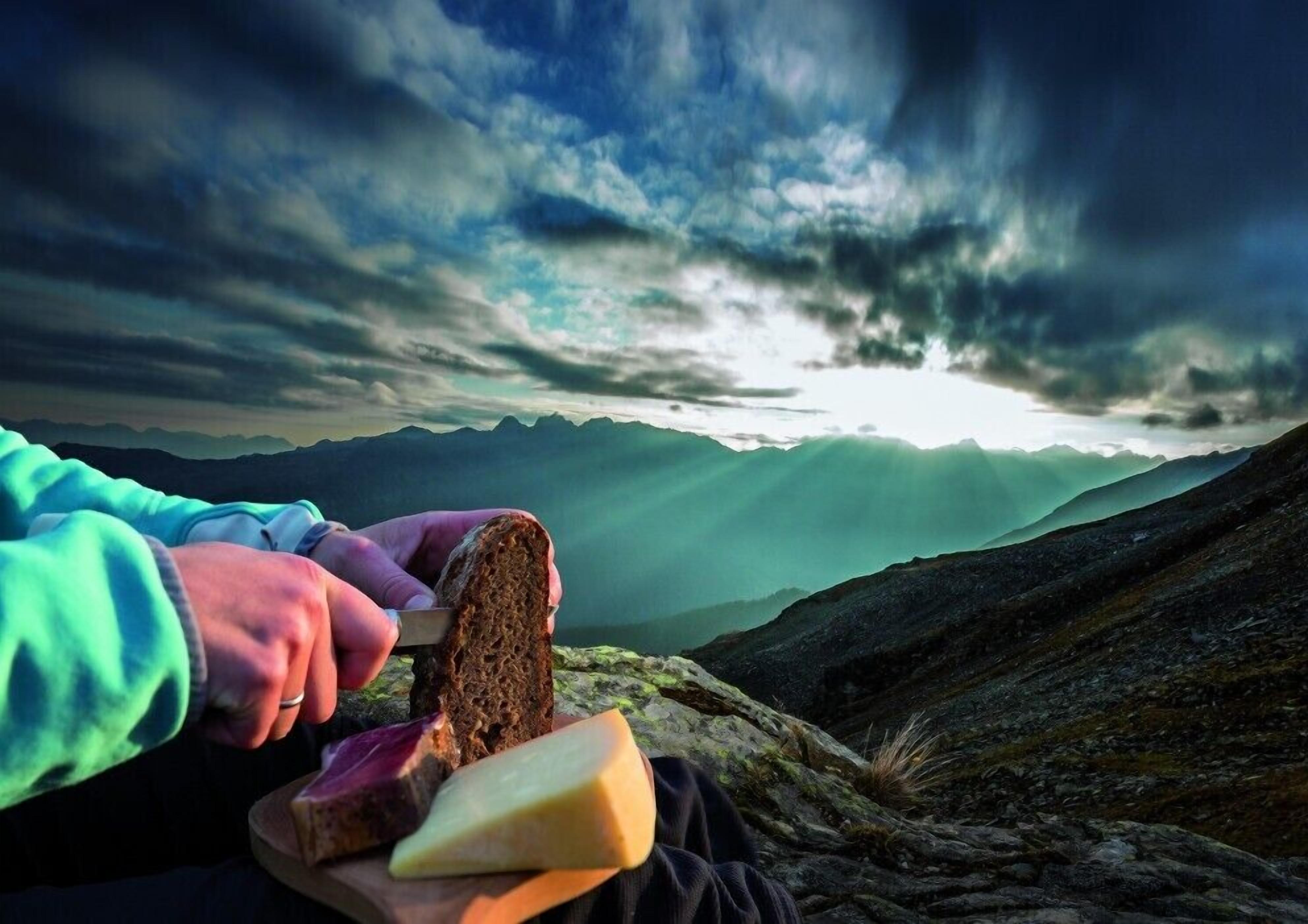 A person holding a slice of bread with a knife, with mountains and a cloudy sky in the background.