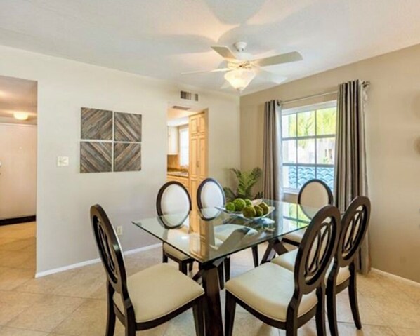 Un coin repas avec une table en verre et quatre chaises, un ventilateur de plafond et une fenêtre avec des rideaux.