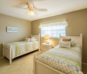 A bedroom with two single beds, a ceiling fan, a bedside table with a lamp, and a window with blinds.
