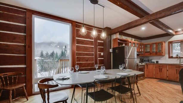 L’intérieur d’une cabane en rondins comprend un coin repas, une cuisine et de grandes fenêtres donnant sur un paysage enneigé.