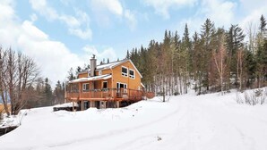 Une maison à deux étages avec un balcon, entourée de neige et d’arbres.