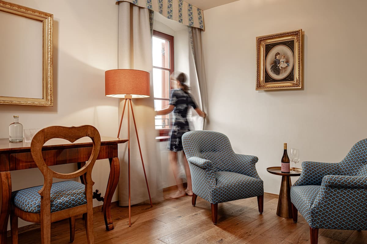 A room with a wooden desk, a chair, a floor lamp, two patterned armchairs, a small table with a bottle and glass, and a framed picture on the wall.