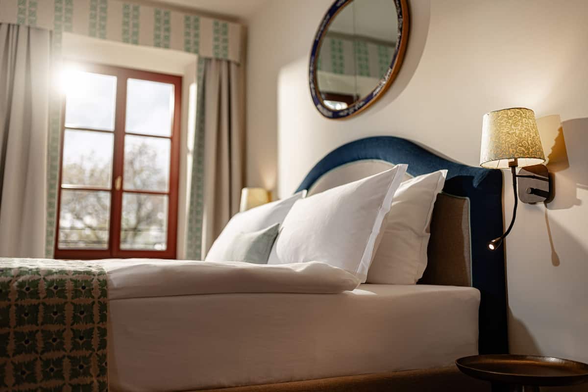 A neatly made bed with white and blue bedding, a round mirror, and a wall-mounted lamp.