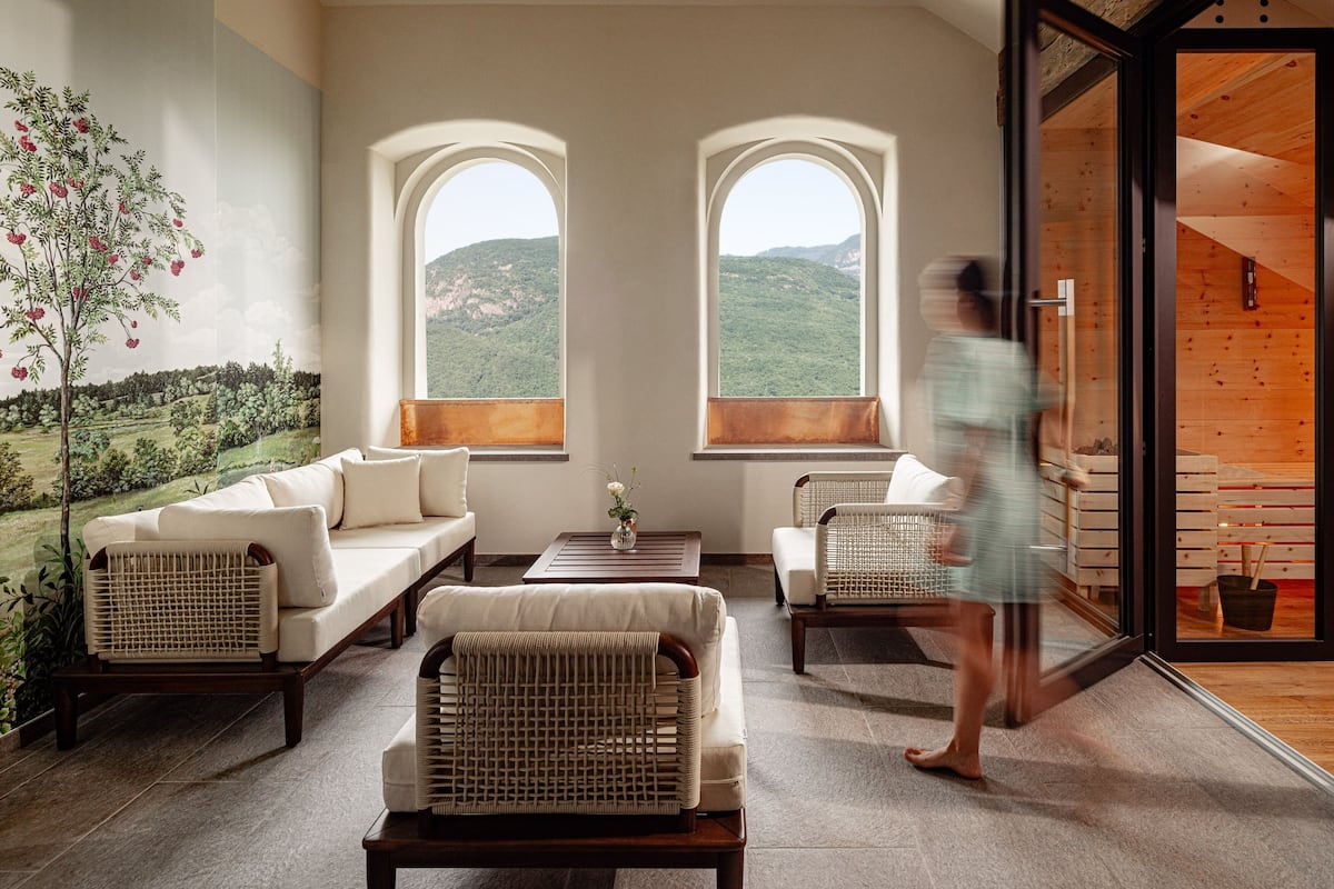 A room with a sofa, chairs, and a table. There are two windows with a view of hills.
