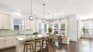 A spacious kitchen with a central island, wooden bar stools, pendant lighting, and large windows providing natural light.