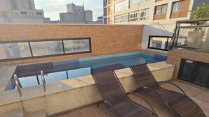 Un espace piscine aménagé sur le toit, avec deux chaises longues, une balustrade en verre et une vue sur le paysage urbain.