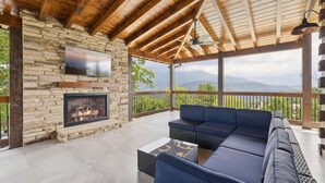 A covered outdoor seating area with a stone fireplace, a large TV mounted on the wall, and a view of mountains.
