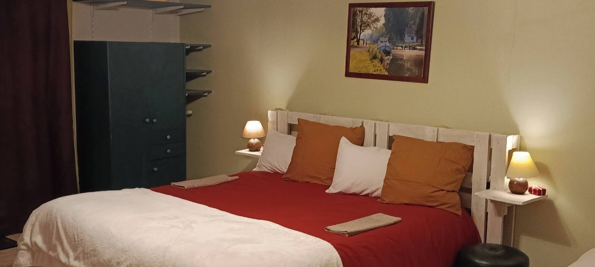 A neatly made bed with a red bedspread and white pillows, flanked by two bedside tables with lamps, a large blue wardrobe, and a painting on the wall.