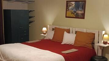 A neatly made bed with a red bedspread and white pillows, flanked by two bedside tables with lamps, a large blue wardrobe, and a painting on the wall.