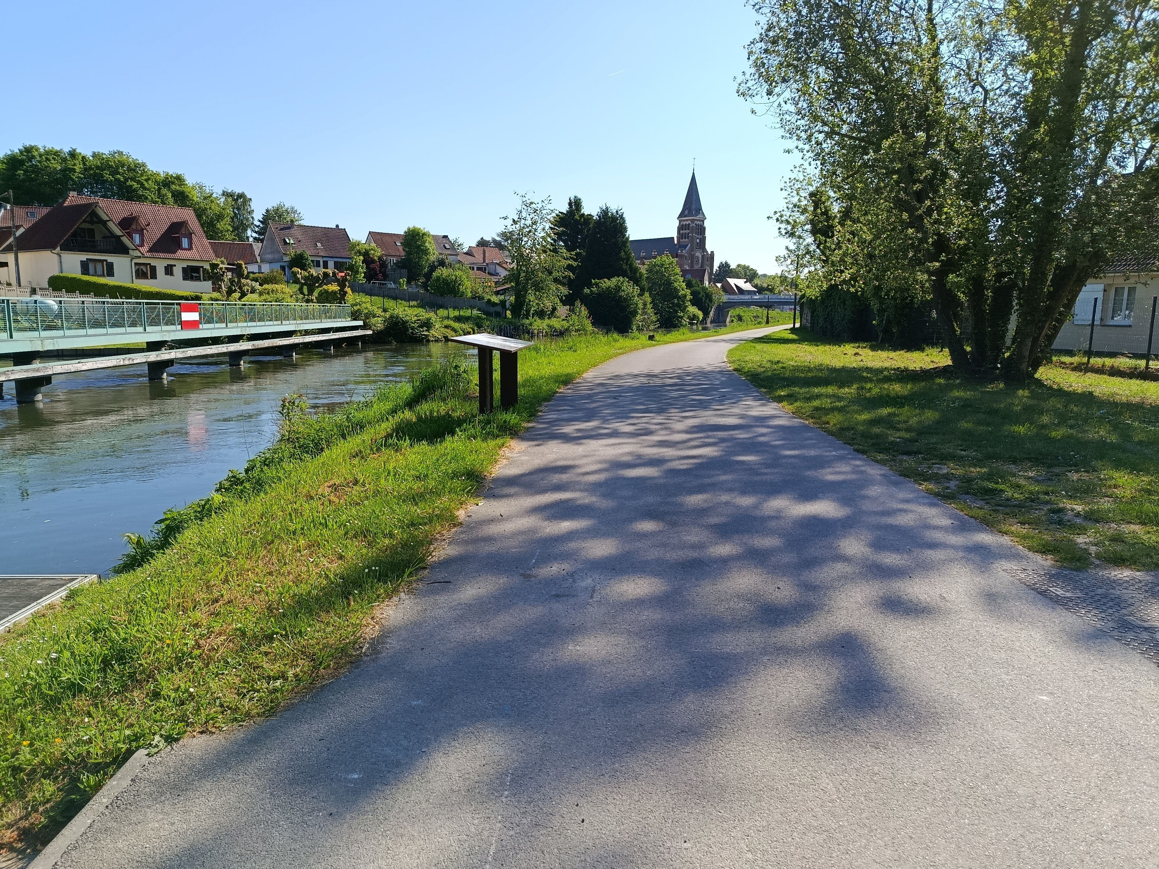 Un sentier pavé longe une rivière, avec un pont, des maisons et une église en arrière-plan.