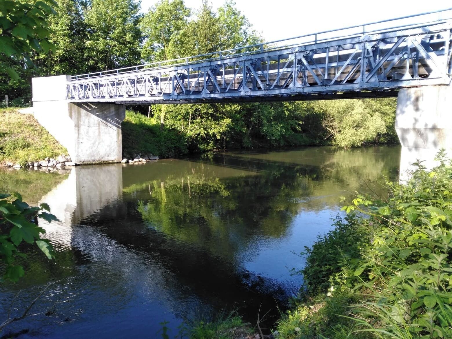 Een metalen brug over een rustige rivier, omzoomd door groen aan weerszijden.