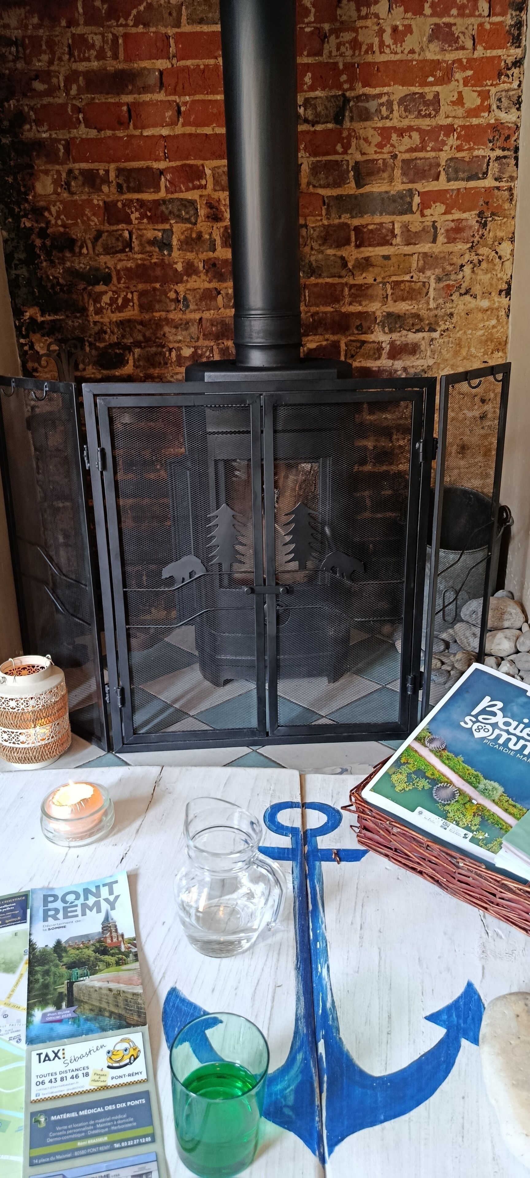 Une cheminée avec une hotte, une table sur laquelle se trouvent divers objets, notamment une bougie, un verre, un livre et un objet décoratif orné d’une ancre bleue.