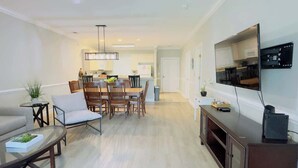 A modern living room with a sofa, coffee table, and TV. There is a dining area with chairs and a table. The kitchen is visible in the background. - Magnificent 2BR condo, ground floor , Pool (Myrtle Beach)