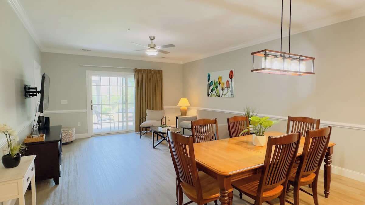 A spacious dining area with a wooden table and chairs, a ceiling fan, and a large window with curtains.
