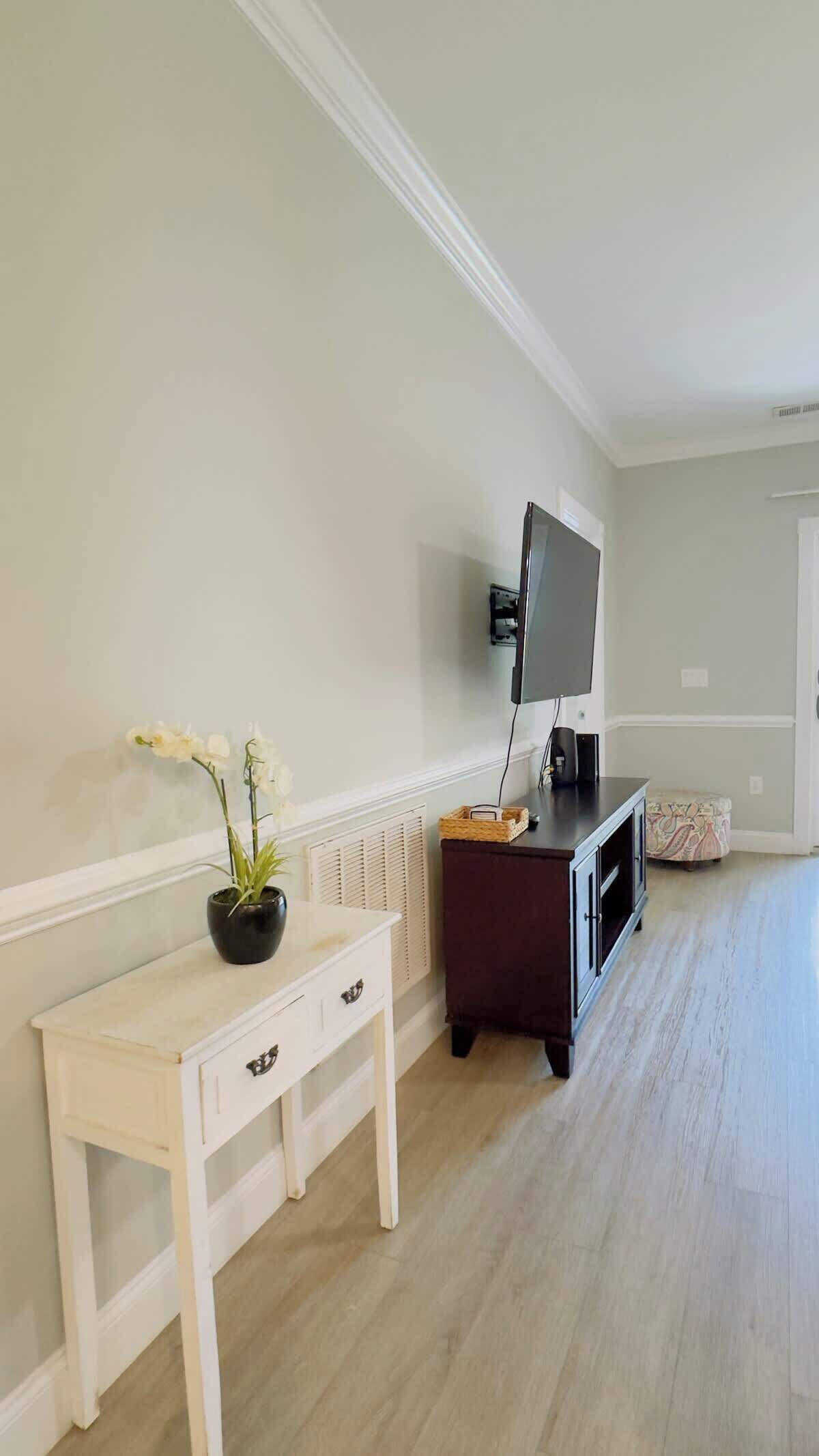 A room with a white console table, a dark wooden cabinet, a flat-screen TV mounted on the wall, and a small white chair.