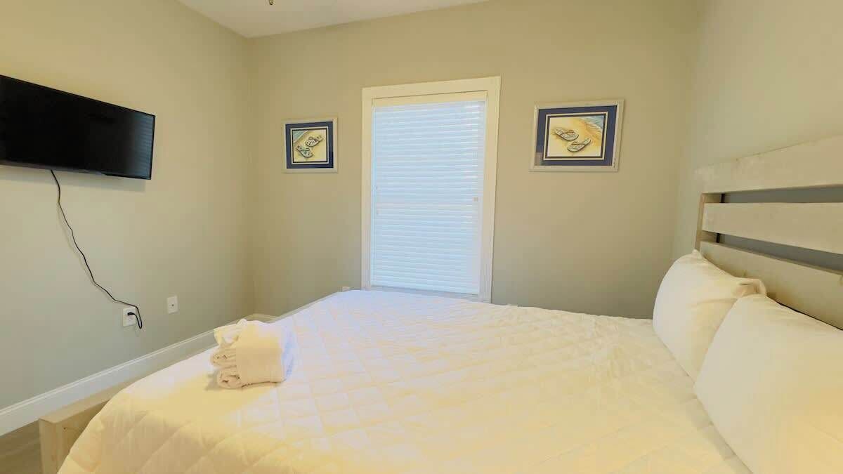 A bedroom with a bed, a window with blinds, a flat-screen TV mounted on the wall, and two framed pictures on the wall.