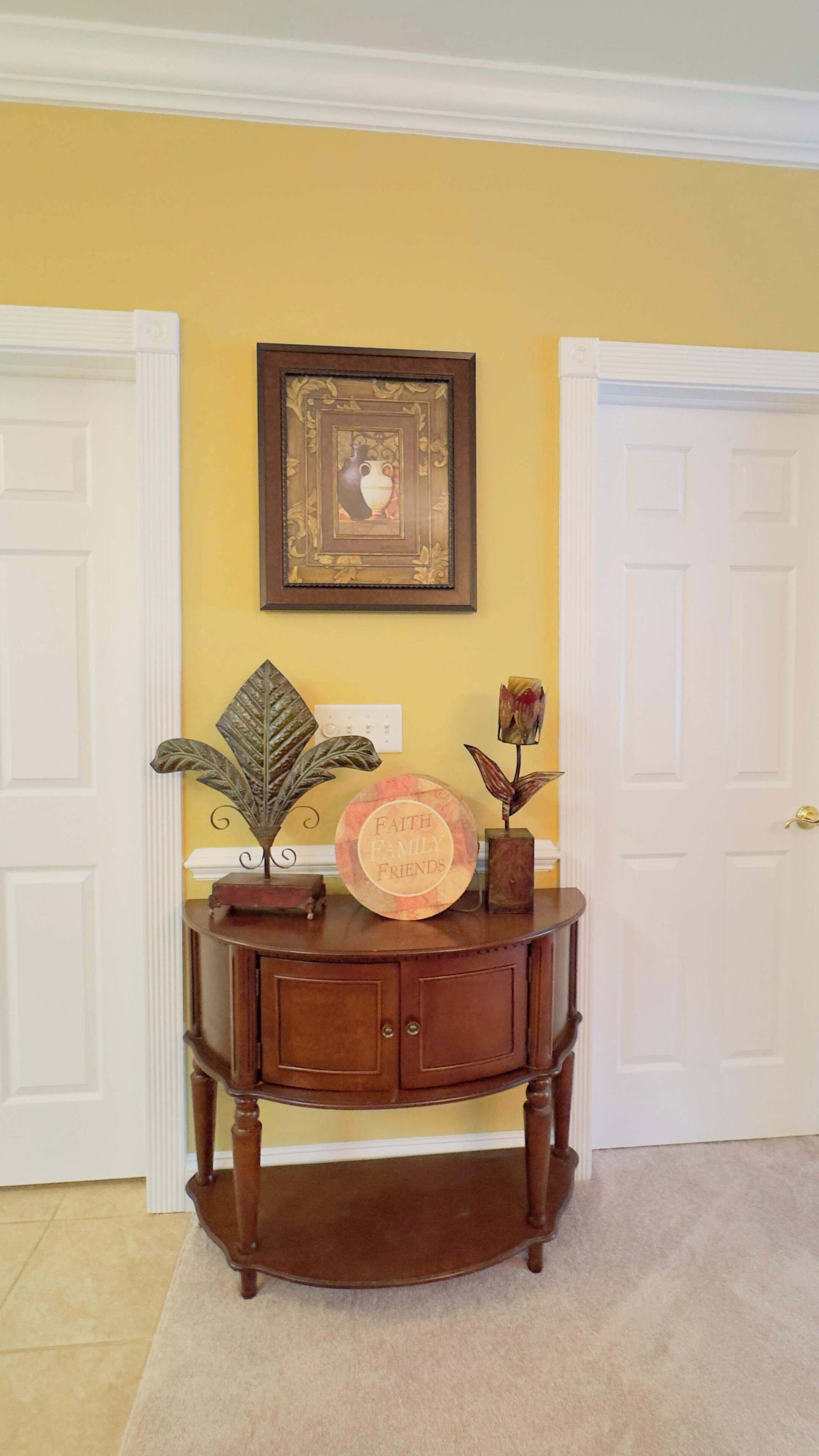 Een houten console tafel met een decoratief blad, een vaas met een bloem, en een bordje met daarop 'FAITH FAMILY FRIENDS' in een kamer met gele muren en witte deuren.